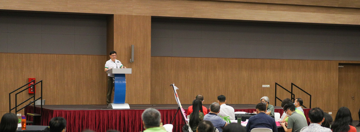 Man speaking at a podium on stage to audience in room with wood paneling.